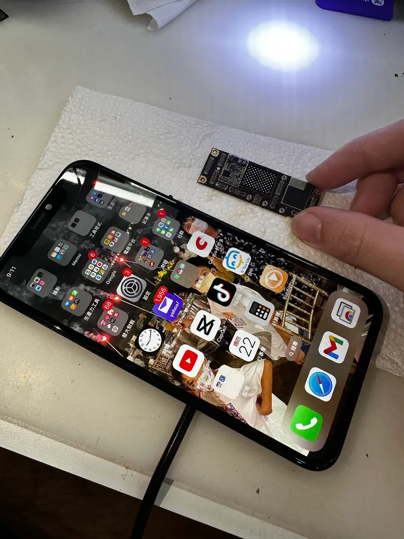 手持修復後的 iPhone 11 顯示正常桌面，桌面上放著維修用的主機板與晶片